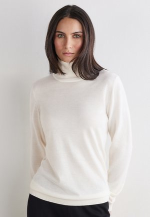 Femme aux cheveux bruns mi-longs, portant un pull à col roulé blanc et un pantalon noir, se tenant devant un mur blanc uni.
