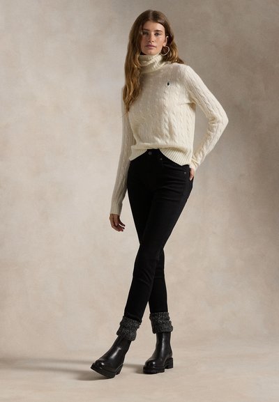 Femme porte un pull à col roulé crème tricoté en torsades, un pantalon noir ajusté, des guêtres grises et des bottines en cuir noir, debout la main dans la poche.