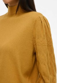Kvinna iklädd en senapsgul stickad tröja med en hög krage och texturerade pösiga ärmar med ett diamantmönster.