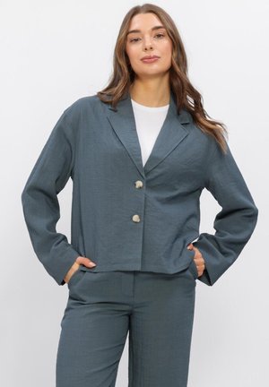 Femme aux longs cheveux bruns portant un blazer bleu-gris à deux boutons et un pantalon assorti, posant les mains dans les poches devant un fond blanc.