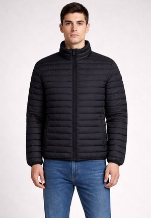 Jonge man die een zwart doorgestikt jack met rits en blauwe spijkerbroek draagt, staand tegen een effen lichte achtergrond.