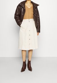 Veste matelassée en cuir marron, haut côtelé beige, jupe en jean blanche trapèze avec boutons sur le devant et bottines marron pointues.