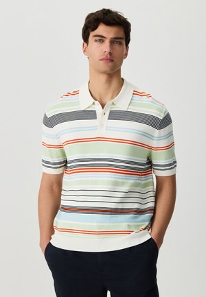 Mladý muž s krátkymi tmavými vlasmi, oblečený v viacfarebnom pruhovanom krátkoročnom polotričku a tmavých nohaviciach, ruky v vreckách, neutrálne pozadie.