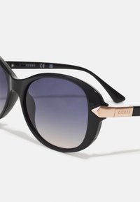 Svart, oversized solbriller med gradientlinser. Stangen har en roségullaccent med "GUESS" gravert. Glatt plastramme.