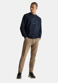 Felpa blu navy con la scritta "est. 1995 LUJO" in un testo più chiaro, abbinata a pantaloni beige e scarpe da ginnastica nere. Vestibilità casual con polsini a coste.