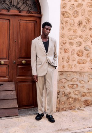 Uomo con abito beige e scarpe nere in piedi con una mano in tasca accanto a una porta di legno e un muro di pietra su una strada lastricata.