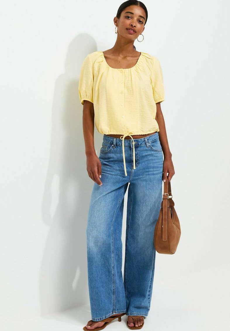 Woman wearing a yellow short-sleeve blouse, wide-leg blue jeans, sandals, hoop earrings, and holding a brown handbag.