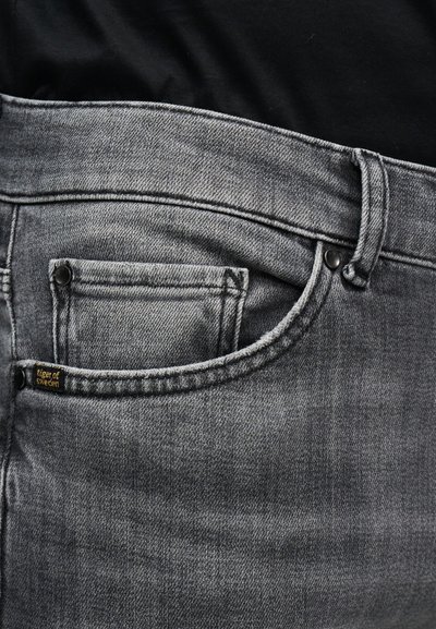 Κοντινό πλάνο τσέπης γκρι τζιν με μαύρα καρφιά και μικρή ετικέτα "tiger of Sweden" σε φόντο μαύρου πουκάμισου.