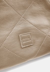 Sac en cuir beige matelassé avec une étiquette rectangulaire embossée portant le texte "FRED'S BRUDER", mettant en valeur le matériau texturé et les coutures.