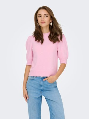 Femme aux longs cheveux bruns portant des boucles d'oreilles argentées en anneau, un pull à manches courtes rose avec de petites perles et un jean bleu clair, une main dans la poche.