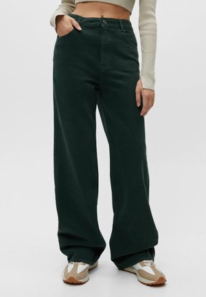 Wide-leg trousers in dark green cotton with a high waist, front pockets, and a single button closure, styled with neutral sneakers.