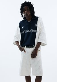 Polo blu navy con testo "Ne Rien Regrette", abbinato a pantaloni corti bianchi testurizzati e una giacca aperta abbinata; gioielli in argento completano il look.