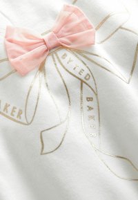 Balts audums T-krekls ar rozā sieta loku un zeltainiem printētiem lentēm ar tekstu "BY TED BAKER." Měksta struktūra, ikdienišķs dizains.