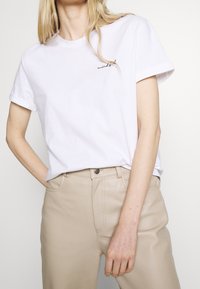 Vit bomull t-shirt med uppvikt ärm, med svart kursiv text "mindful" på vänster bröst, matchat med ljusbeige byxor.