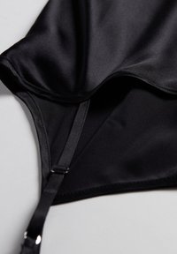 Tejido de satén negro, textura suave, diseño de lencería con tirantes ajustables, costuras laterales y una silueta elegante, enfocado en la cobertura y el ajuste.