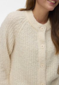 Frau trägt eine cremefarbene, flauschige Strickjacke mit großen Knöpfen und langen Ärmeln, lächelt sanft.