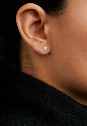 Záber na ucho s malou, okrúhlou, textúrovanou striebristou náušnicou na pozadí tmavých vlasov a čiernej látky.