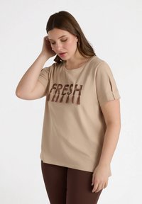 Camiseta de manga corta beige con el texto bordado "FRESH" en marrón, que presenta un cuello redondo y acentos de manga enrollada. Textura de tejido suave.