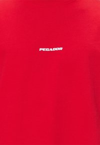 Punainen puuvillainen t-paita pyöreällä kaula-aukkolla. Rinnassa valkoinen "PEGADOR"-logo. Sileä pinta ja minimalistinen design.