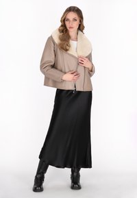 Giacca in pelle beige con un ampio colletto di pelliccia, chiusura con zip sul davanti, abbinata a una lunga gonna di raso nera e stivali neri sopra il ginocchio.