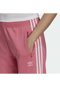 Růžové běžecké kalhoty Adidas s bílými postranními pruhy, elastickým pasem a bočními kapsami. Na stehně se nachází logo. Hladká textilie.
