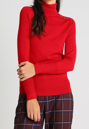 Femme portant un pull à col roulé rouge avec des détails cloutés aux épaules et un pantalon marron à carreaux, les bras croisés à la taille.