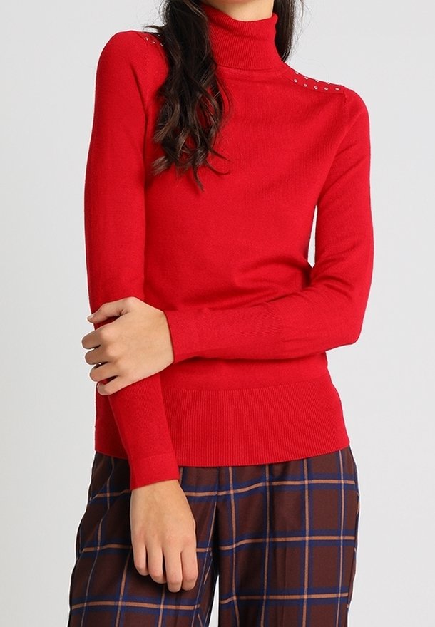 Femme portant un pull à col roulé rouge avec des détails cloutés aux épaules et un pantalon marron à carreaux, les bras croisés à la taille.