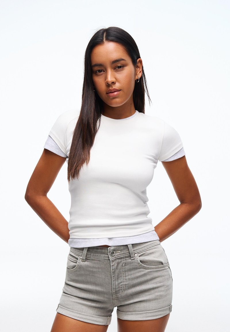 Jeune femme aux longs cheveux foncés portant un t-shirt blanc ajusté et un short en denim gris clair, debout les mains derrière le dos.