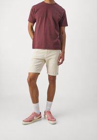 Muž v vínovém tričku, světle béžových krátkých kalhotách na rolování, bílých ponožkách do poloviny lýtek a růžových teniskách s bílým znakem a krémovými podrážkami.