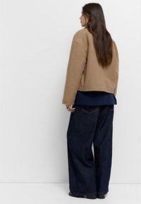 Brun bomuldsjakke, afslappet pasform, med en kant ved taljen. Parret med en marineblå top og mørk denim brede bukser. Langt, lige hår synligt.