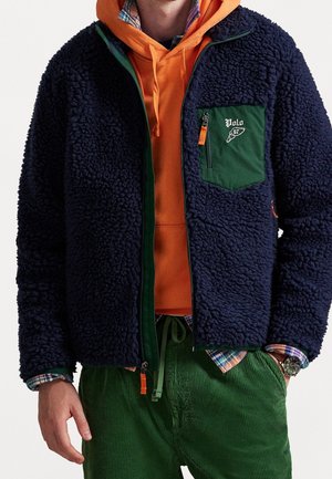 Hombre con chaqueta de forro polar azul marino con bolsillo y cremallera verdes sobre sudadera naranja, camisa de cuadros y pantalones de pana verdes.