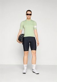 Lichtgroen fietsshirt met korte mouwen, zwarte gewatteerde shorts en witte fietsschoenen. De fietser houdt een zwarte helm vast en draagt een zonnebril.