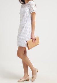 White perforated dress with short sleeves, beige flat shoes with a bow detail, and a matching beige clutch handbag.