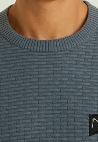 Gray textured knit sweater featuring a ribbed crew neck and a small black logo patch on the left side.