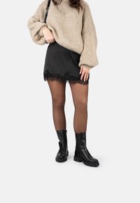 Minifalda de satén negra con encaje, suéter de punto beige oversize, botines negros y medias transparentes. Diseño de outfit simple y en capas.