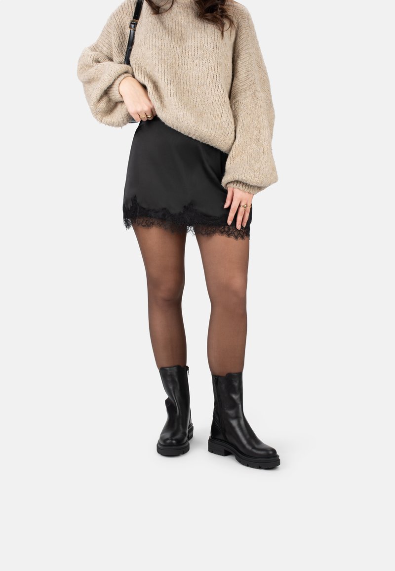 Minifalda de satén negra con encaje, suéter de punto beige oversize, botines negros y medias transparentes. Diseño de outfit simple y en capas.