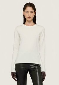 Bílá košile s dlouhým rukávem z měkké látky, s kulatým výstřihem a přiléhavým střihem. Nošena s černými koženými kalhotami a tmavými rukavicemi.