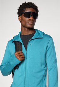 Turkizna jakna na zadrgo s kapuco, dolgimi rokavi in odprtinami za palce, nosi se čez črno majico. Model drži trak črnega nahrbtnika.