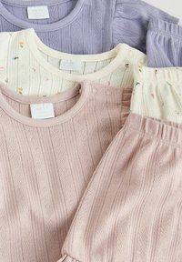 Trois ensembles de layette tricotée en violet, crème avec imprimé floral, et rose, comprenant des hauts avec boutons et des pantalons assortis en côtes.
