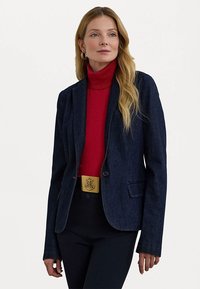 Temno modra blazer s strukturirano zasnovo, zapiranjem na en gumb in z izrezanimi reverji, v kombinaciji s rdečim puloverjem.