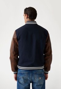 Chaqueta bomber navy con mangas de cuero marrón, cuello acanalado a rayas y puños. Llevada sobre unos vaqueros de mezclilla azules, mostrando la vista trasera.