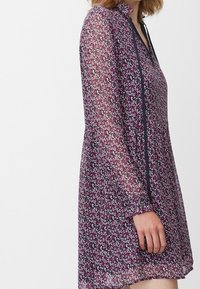 Lila blommig klänning i genomskinligt tyg med långa ärmar, utrustad med en krage och mörk kant längs fram.