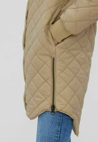 Veste matelassée beige avec une fermeture éclair sur le côté, des poignets côtelés et un ourlet anglé, présentant une texture douce et lisse ainsi qu'un motif en losanges.