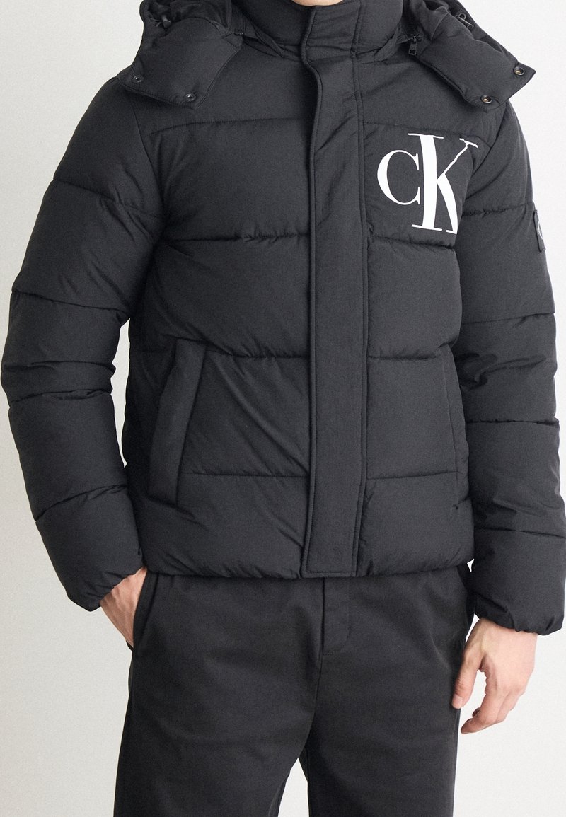 Veste matelassée noire avec un col montant, une capuche et une fermeture éclair à l'avant. Présente un grand logo blanc "CK" sur le côté gauche de la poitrine et des coutures horizontales.