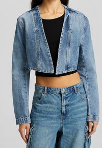 Zkrácená džínová bunda v světle modré barvě se štěpením vepředu, dlouhými rukávy a viditelnými švy. Nositá přes černý ribbovaný top.