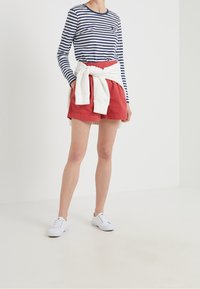 Maglietta a maniche lunghe a righe blu navy e bianche abbinata a pantaloncini rossi. Felpa bianca legata in vita e scarpe da ginnastica bianche ai piedi.