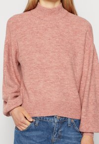 Ljust rosa ribbad tröja med hög krage och vida ärmar, tillverkad av mjukt stickat material. Matchas med blå denimjeans.