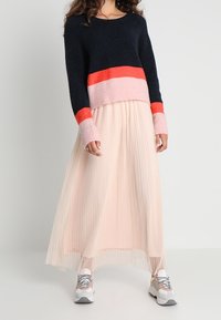 Pull en maille à rayures bleu marine et rose avec une jupe plissée rose pâle et des baskets grises. Les matériaux semblent doux, avec un design en couches et fluide.