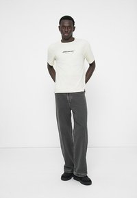Ung mann står med hendene bak ryggen iført hvit McLaren Formel 1 t-skjorte, løse grå jeans og sorte sko foran en ensfarget bakgrunn.