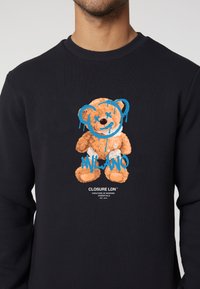 Black sweatshirt featuring a graphic of a bear with a blue smiley face, accompanied by the text "MILANO" in blue and brand details below.